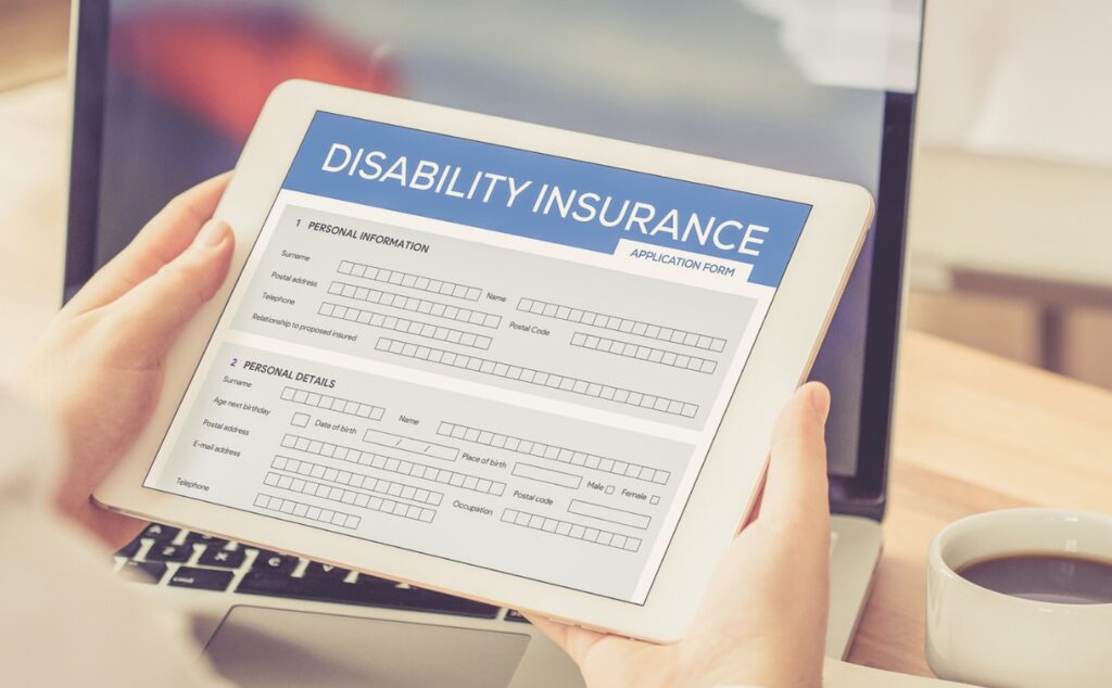 A pair of hands holds an electronic tablet with a disability insurance application form on its screen, ideally a disability insurance application for own specialty disability insurance for physicians. The words DISABILITY INSURANCE is at the top of the page with two sections, “Personal Information” and “Personal Details”. Each section has blank boxes in which to put information such as your name, age, address and telephone number.