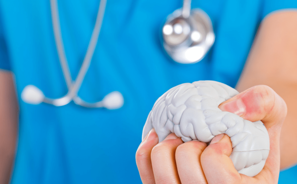 A female doctor’s hand holds a facsimile of a brain in her fist, squeezing it to signify how mental health issues affect medical residents. She wears scrubs and has a stethoscope around her neck.